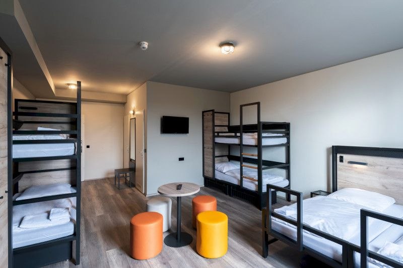 Ein modernes Familienzimmer mit zwei Etagenbetten, einem Einzelbett, einem kleinen Tisch und bunten Hockern. An der Wand hängt ein Fernseher., © A&O Hostels Marketing GmbH