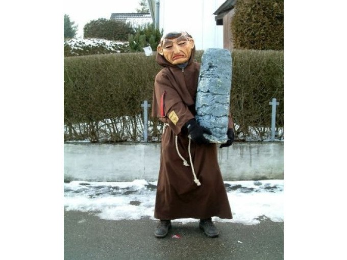 Person in braunem Kost&uuml;m mit Maske h&auml;lt einen gro&szlig;en grauen Stein. Schnee liegt am Boden, Hecke im Hintergrund., &copy; Cool-Tours StattReisen