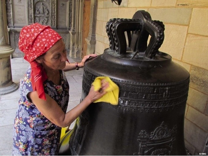 Frau mit rotem Kopftuch poliert eine große Glocke mit einem gelben Tuch in einem historischen Gebäude., © Stuttgart Marketing GmbH Frau mit rotem Kopftuch poliert eine große Glocke mit einem gelben Tuch in einem historischen Gebäude., © Stuttgart Marketing GmbH