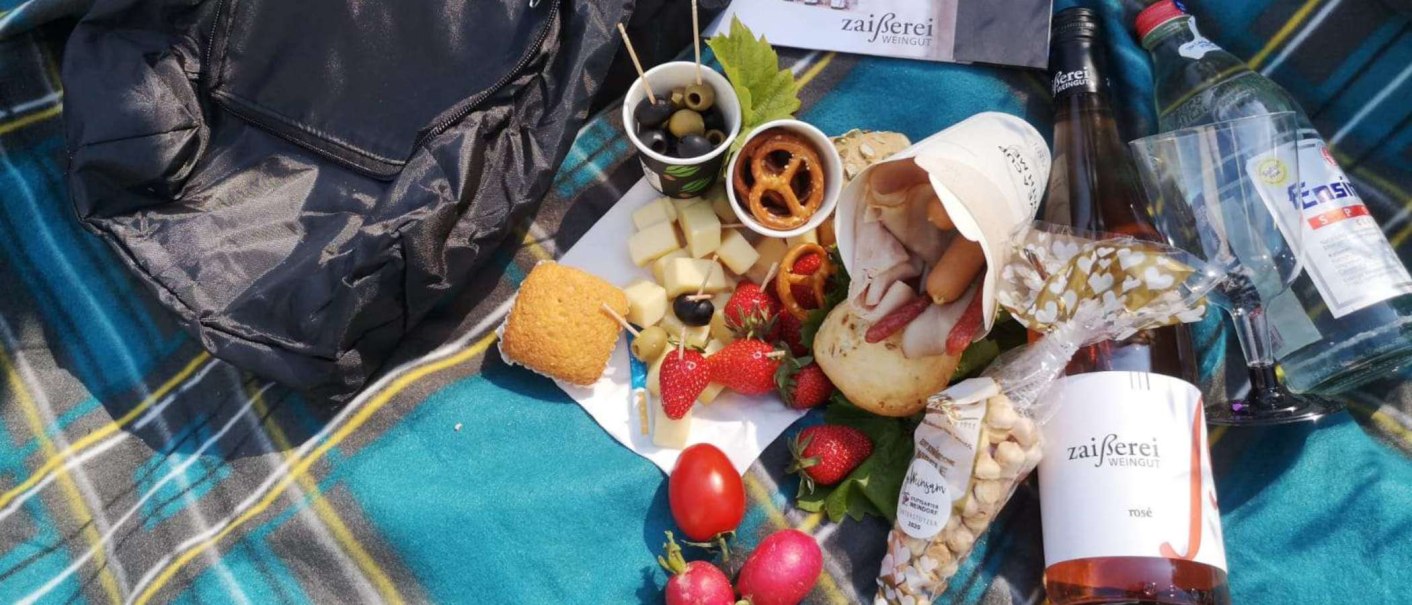 Ein Picknick auf einer karierten Decke mit Käse, Oliven, Erdbeeren, Brot, Wein und Wasserflasche. Ein Rucksack liegt daneben., © Weingut Zaißerei Ein Picknick auf einer karierten Decke mit Käse, Oliven, Erdbeeren, Brot, Wein und Wasserflasche. Ein Rucksack liegt daneben., © Weingut Zaißerei
