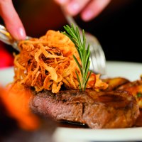 Zwiebelrostbraten mit knusprigen Röstzwiebeln und Rosmarin auf einem Teller, im Hintergrund unscharf gedeckter Tisch., © Fotolia, Kzenon