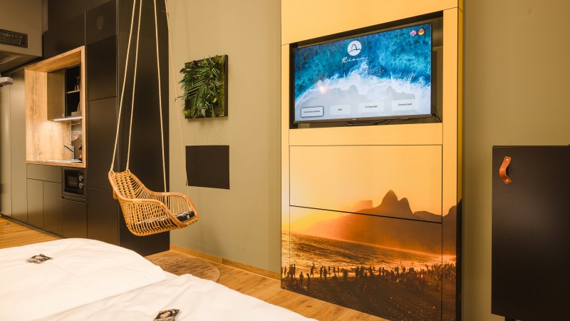 Modernes Hotelzimmer mit Hängesessel, Fernseher mit Strandbild, kleiner Küchenzeile und dekorativem Pflanzenbild an der Wand., © TOMAS