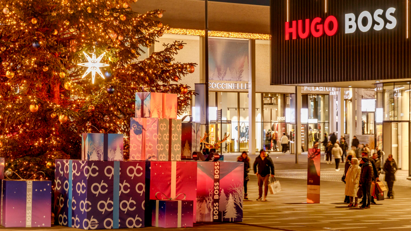 Weihnachtlich geschmückter Platz mit großem Baum und Geschenken vor dem Hugo Boss Geschäft. Menschen flanieren in der festlichen Atmosphäre., © OUTLETCITY AG