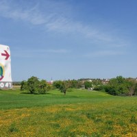 Ein Fahrradweg-Schild an einem Pfosten vor einer gr&uuml;nen Wiese mit gelben Blumen. Im Hintergrund ist ein Dorf zu sehen., &copy; Natur.Nah. Sch&ouml;nbuch & Heckeng&auml;u