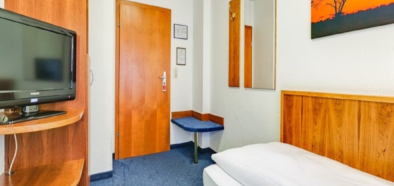 Ein gemütliches Einzelzimmer mit einem Bett, einem Fernseher, Holzmöbeln und einem Bild an der Wand. Der Teppich ist blau., © ARNOTEL GmbH Ein gemütliches Einzelzimmer mit einem Bett, einem Fernseher, Holzmöbeln und einem Bild an der Wand. Der Teppich ist blau., © ARNOTEL GmbH