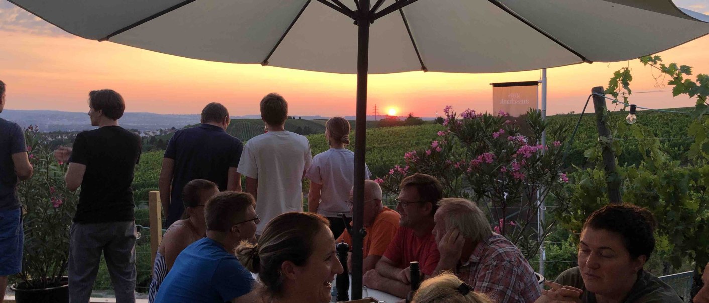Menschen sitzen unter Sonnenschirmen in einem Weinberg und genießen den Sonnenuntergang. Einige stehen und blicken in die Ferne., © Weingut Diehl