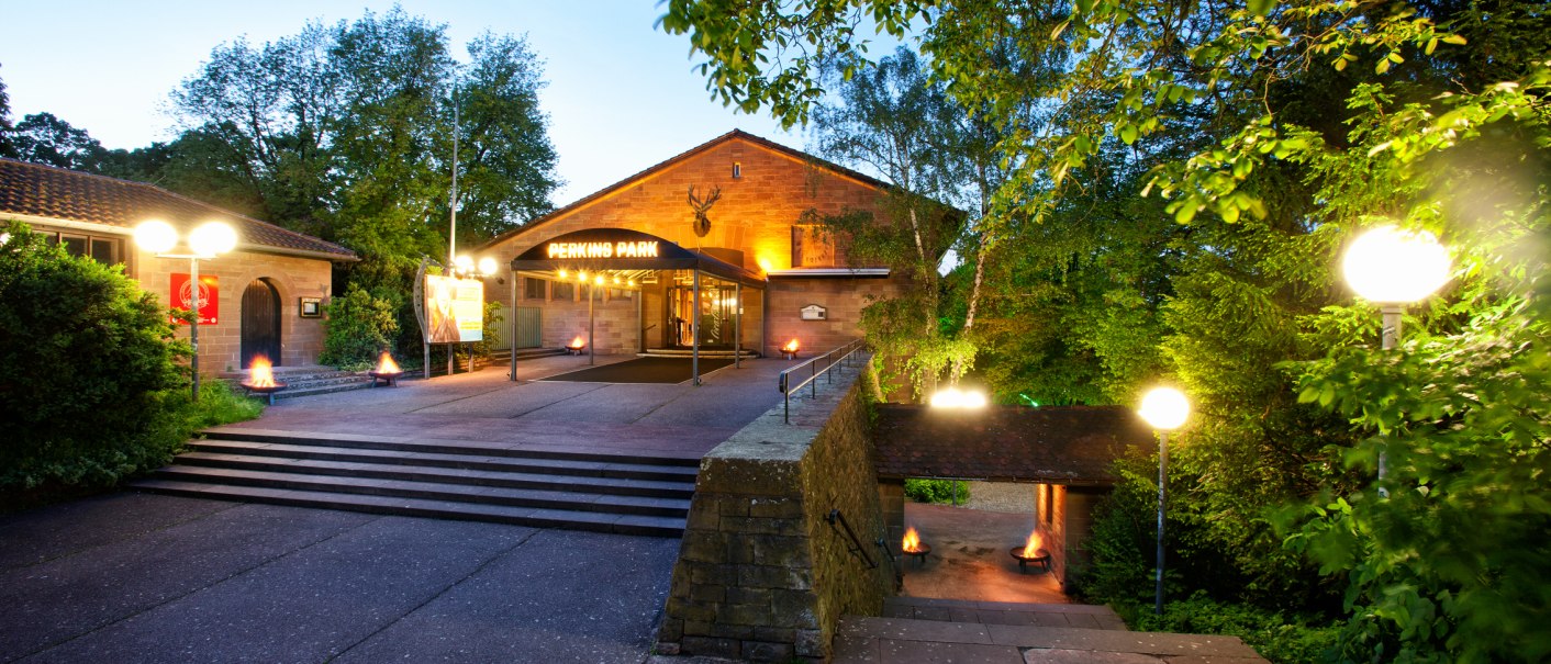 Eingang des Perkins Park in Stuttgart bei Dämmerung, umgeben von Bäumen und beleuchtet mit Laternen., © PerkinsPark Eingang des Perkins Park in Stuttgart bei Dämmerung, umgeben von Bäumen und beleuchtet mit Laternen., © PerkinsPark