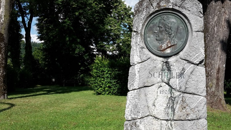 Schillerdenkmal in gr&uuml;ner Parkanlage mit B&uuml;schen und B&auml;umen - grob behauene, graue Natursteins&auml;ule, mit bronzenem Port&auml;t-Medaillon, darunter die Inschrift "Schiller" und die Jahreszahl 1905, &copy; Petra Natzkowski