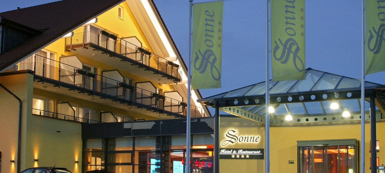 Hotel Sonne bei Dämmerung, beleuchteter Eingang, gelbes Gebäude mit Balkonen, drei Fahnen davor., © Hotel & Restaurant Sonne