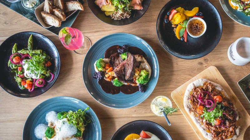 Ein Tisch mit verschiedenen Gerichten: Fleisch mit Gem&uuml;se, Salate, Burger mit Pommes, Brot und Desserts. Getr&auml;nke sind ebenfalls zu sehen., &copy; SatzDrei