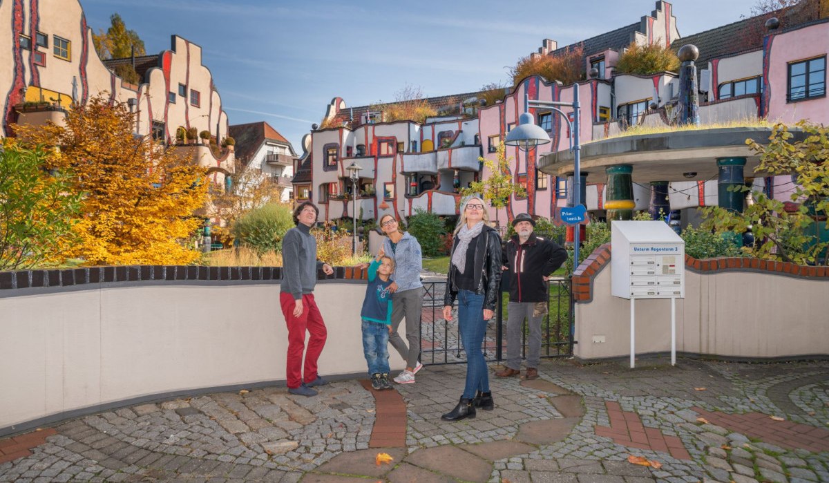 Menschen stehen vor einem bunten Hundertwasser-Gebäude mit unregelmäßigen Formen und begrünten Dächern., © Bildergalerie Attila Menschen stehen vor einem bunten Hundertwasser-Gebäude mit unregelmäßigen Formen und begrünten Dächern., © Bildergalerie Attila