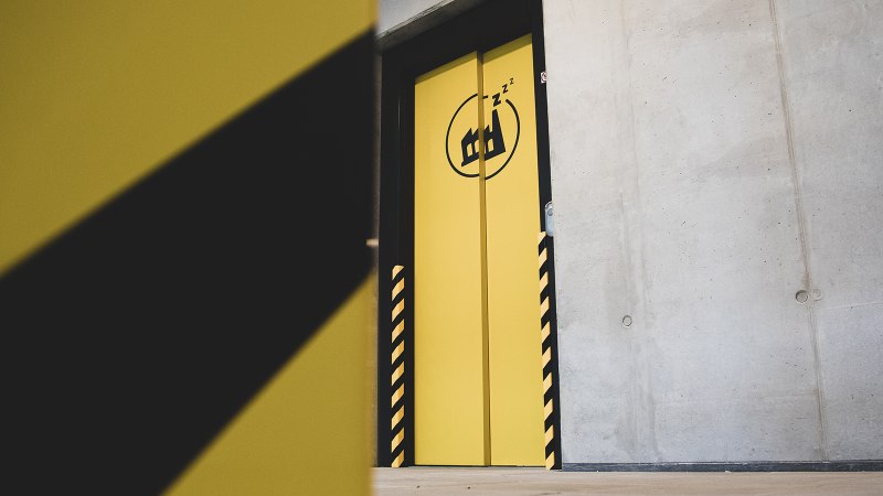 Gelbe Tür mit schwarzem Logo und Zzz-Symbol in einem Betonraum. Die Tür hat schwarz-gelbe Streifen an den Seiten., © Das Schlafwerk - DQuadrat Living GmbH