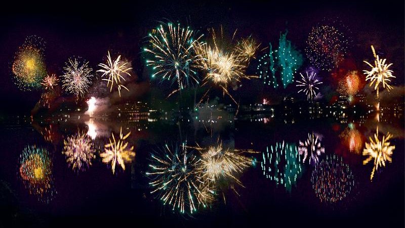 Ein farbenfrohes Feuerwerk erleuchtet den Nachthimmel über einem See, dessen Wasser die Lichter reflektiert. Die Stadtlichter sind im Hintergrund sichtbar., © Backnang Ein farbenfrohes Feuerwerk erleuchtet den Nachthimmel über einem See, dessen Wasser die Lichter reflektiert. Die Stadtlichter sind im Hintergrund sichtbar., © Backnang
