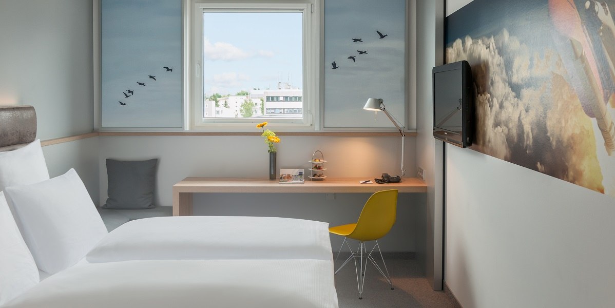 Modernes Doppelzimmer mit weißem Bett, Schreibtisch, gelbem Stuhl und Fenster mit Stadtblick. Wanddekoration mit Vogelmotiv., © Essential by Dorint