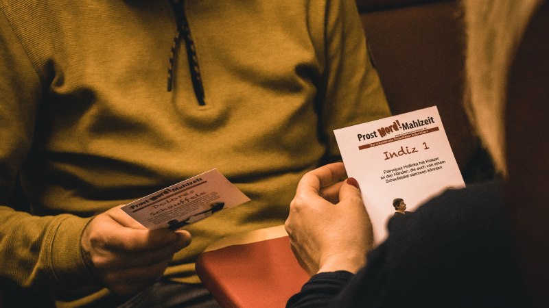 Zwei Personen lesen Karten mit Hinweisen bei einem Krimi-Dinner. Eine Karte trägt den Titel 'Indiz 1'. Es herrscht eine gemütliche Atmosphäre., © Stadt Welzheim
