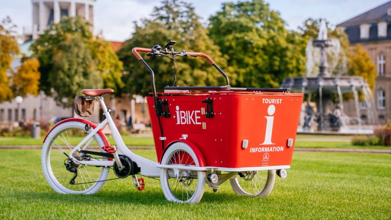 Ein rotes Lastenfahrrad mit der Aufschrift 'I-Bike' steht auf einer Wiese vor einem Brunnen und einem Gebäude mit Kuppel., © Stuttgart-Marketing GmbH, Thomas Niedermüller Ein rotes Lastenfahrrad mit der Aufschrift 'I-Bike' steht auf einer Wiese vor einem Brunnen und einem Gebäude mit Kuppel., © Stuttgart-Marketing GmbH, Thomas Niedermüller