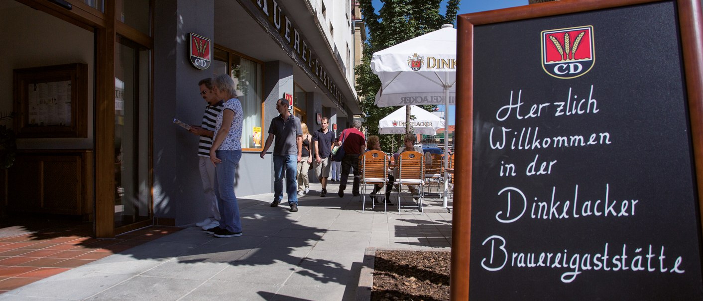 Eingang der Dinkelacker Brauereigaststätte mit einem Willkommensschild. Menschen gehen vorbei, einige sitzen an Tischen unter Sonnenschirmen., © Familienbrauerei Dinkelacker