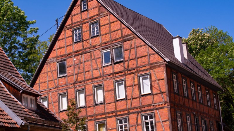 Ein gro&szlig;es Fachwerkhaus mit rotem Ziegelmauerwerk und dunklem Dach steht vor einem klaren blauen Himmel. B&auml;ume umgeben das Geb&auml;ude., &copy; Agentur Arcos