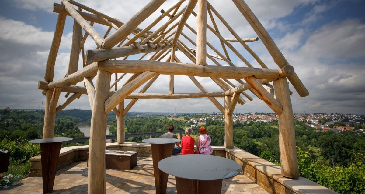 Drei Personen sitzen unter einer hölzernen Pergola mit Blick auf eine grüne Landschaft und eine Stadt im Hintergrund. Der Himmel ist bewölkt., © Stoppel Drei Personen sitzen unter einer hölzernen Pergola mit Blick auf eine grüne Landschaft und eine Stadt im Hintergrund. Der Himmel ist bewölkt., © Stoppel