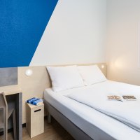 Ein schlichtes Hotelzimmer mit Doppelbett, blau-weißer Wand, kleinem Schreibtisch und Fenster mit Blick ins Grüne., © DQuadrat Living GmbH