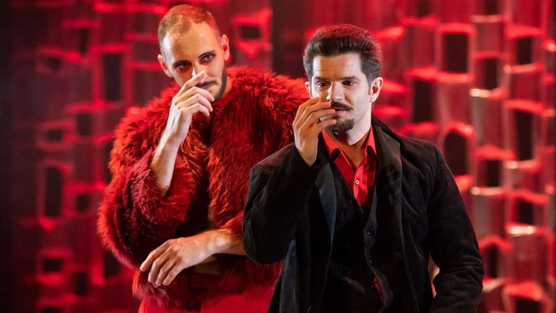 Zwei Männer in auffälliger Kleidung stehen auf einer Bühne. Der Hintergrund ist rot beleuchtet, was eine dramatische Atmosphäre schafft.