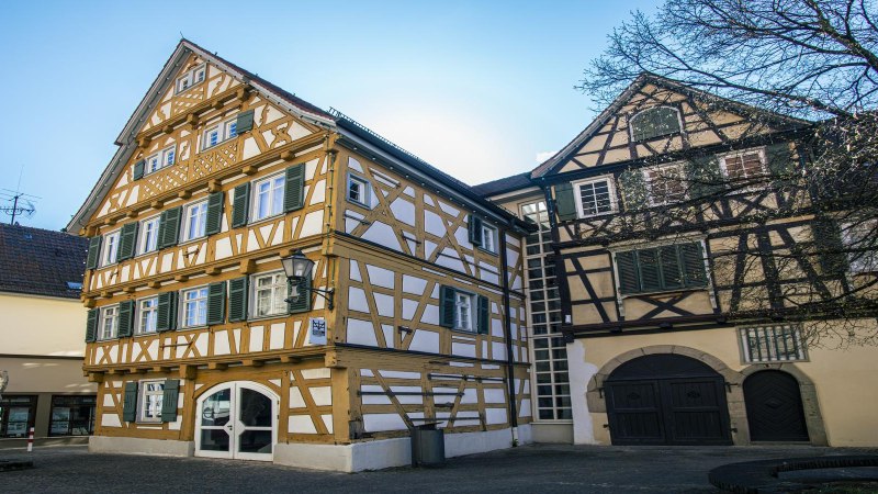 Fachwerkhäuser in Schorndorf, darunter das Stadtmuseum, bei sonnigem Wetter. Ein Baum und eine Skulptur sind ebenfalls zu sehen., © SMG, Sarah Schmid