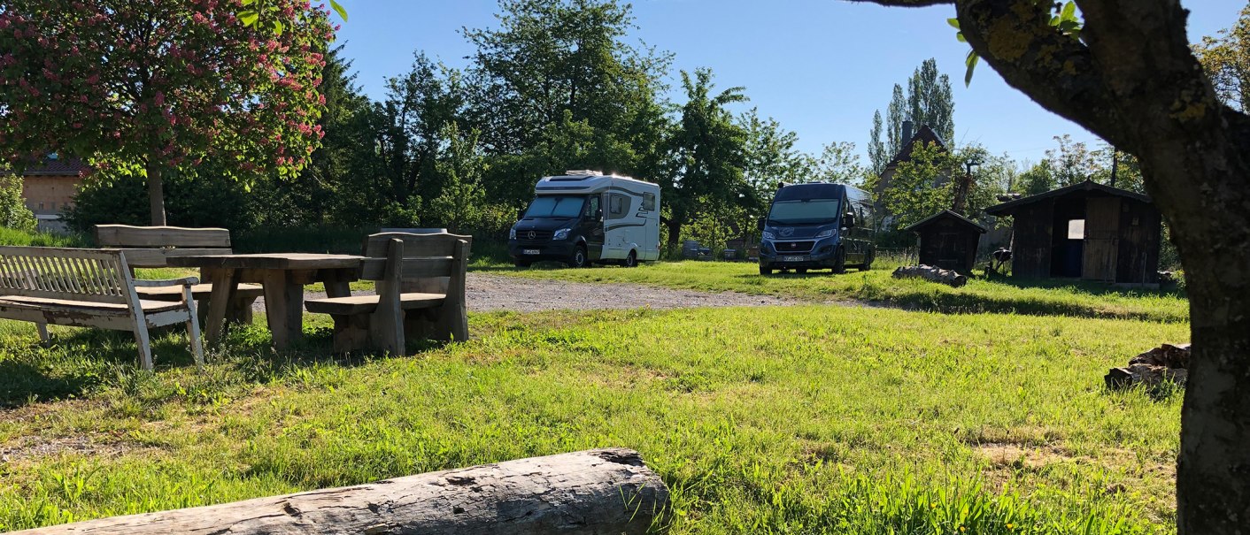Wohnmobilstellplatz mit zwei Wohnmobilen, Holztisch und Bänken unter einem blühenden Baum. Im Hintergrund sind kleine Holzhütten zu sehen., © Bio Weingut Zimmer