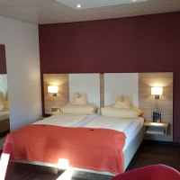 Modernes Doppelzimmer mit zwei Betten, roter Wand, Fernseher und stilvoller Beleuchtung. Ein Tisch mit Wasserflasche und Gl&auml;sern steht bereit., &copy; Hotel Koch