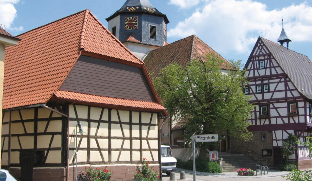 Fachwerkhäuser in Gündelbachs Ortsmitte, mit einem Kirchturm im Hintergrund. Ein Straßenschild zeigt 'Winzerstraße'. Sonniger Tag mit blauem Himmel., © Land der 1000 Hügel - Kraichgau-Stromberg Fachwerkhäuser in Gündelbachs Ortsmitte, mit einem Kirchturm im Hintergrund. Ein Straßenschild zeigt 'Winzerstraße'. Sonniger Tag mit blauem Himmel., © Land der 1000 Hügel - Kraichgau-Stromberg