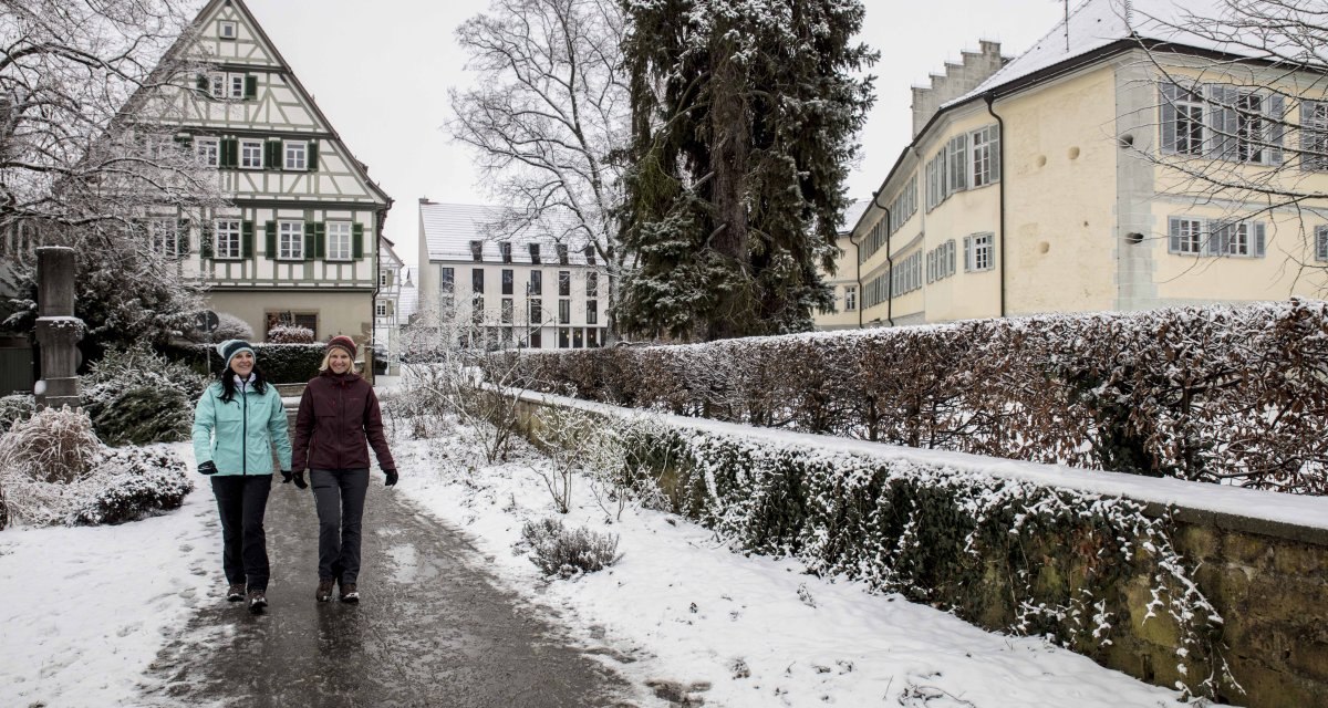 Zwei Personen spazieren auf einem verschneiten Weg in einer winterlichen Stadt. Im Hintergrund sind ein Fachwerkhaus und schneebedeckte Bäume zu sehen., © Schwäbische Alb Tourismusverband e.V. Zwei Personen spazieren auf einem verschneiten Weg in einer winterlichen Stadt. Im Hintergrund sind ein Fachwerkhaus und schneebedeckte Bäume zu sehen., © Schwäbische Alb Tourismusverband e.V.