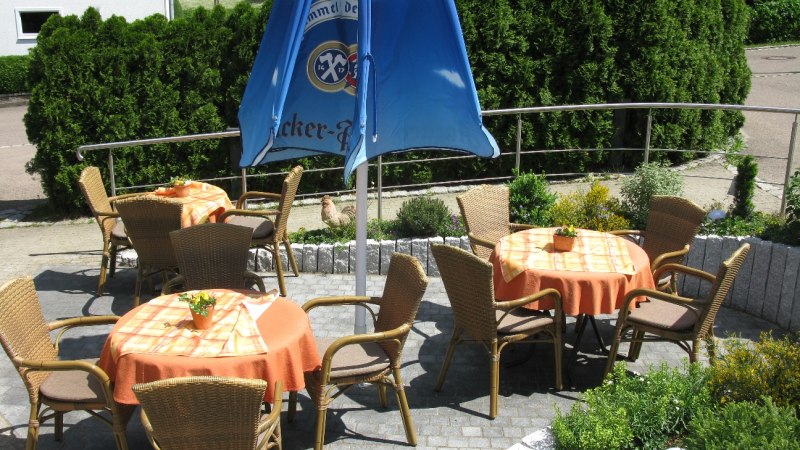 Terrasse mit mehreren Tischen und Stühlen, orange Tischdecken, blauer Sonnenschirm, umgeben von grünen Pflanzen., © Freimut Hößle