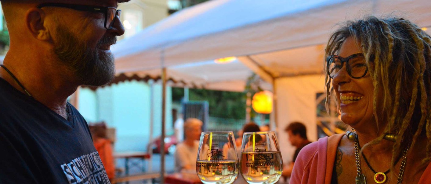 Zwei Personen stoßen mit Weingläsern an und lächeln sich an, während sie unter einem Zelt sitzen., © Weingut Ruoff Zwei Personen stoßen mit Weingläsern an und lächeln sich an, während sie unter einem Zelt sitzen., © Weingut Ruoff