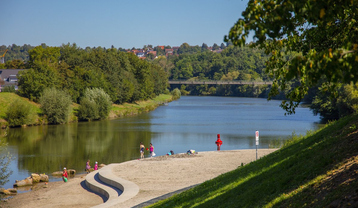Menschen entspannen am sandigen Ufer des Remseckufers, umgeben von grünen Bäumen und einem ruhigen Fluss. Eine Brücke ist im Hintergrund sichtbar., © Remstal Tourismus e.V.