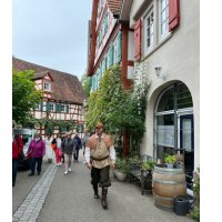 Ein Mann in historischer Kleidung geht durch eine malerische Stra&szlig;e in Schorndorf, flankiert von Fachwerkh&auml;usern und einer Gruppe von Besuchern., &copy; Eigenbetrieb Tourismus und Citymanagement Schorndorf