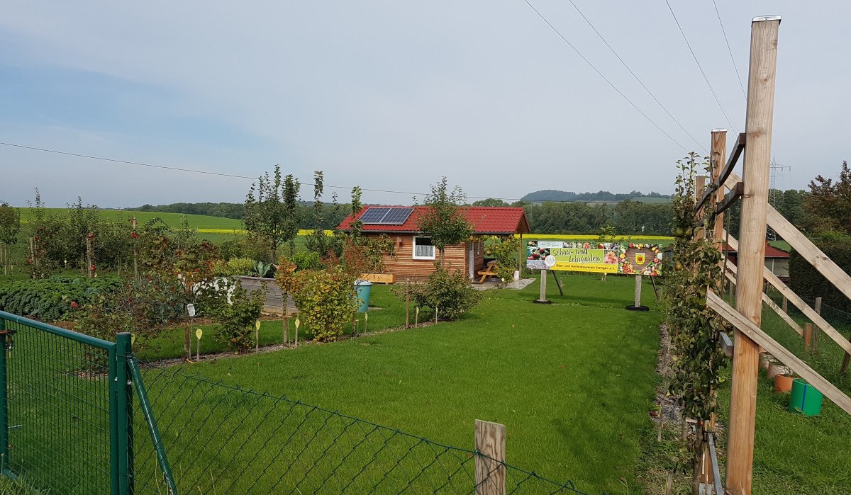 Ein gepflegter Garten mit Rasen, Bäumen und einem Holzhaus. Ein Zaun umgibt das Gelände, im Hintergrund ist ein Banner sichtbar., © Remstal Tourismus e.V.