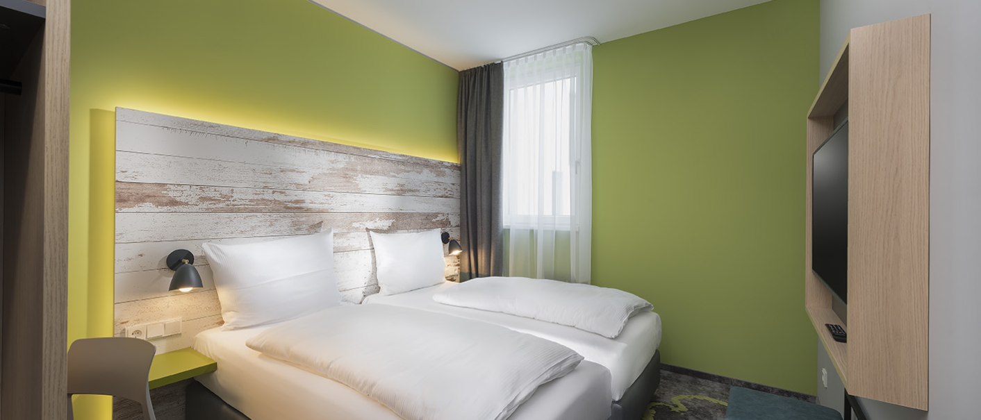 Modernes Hotelzimmer mit zwei Einzelbetten, grüne Wand, Holzkopfteil, Fernseher und Fenster mit Vorhängen., © ibis Styles Stuttgart Vaihingen