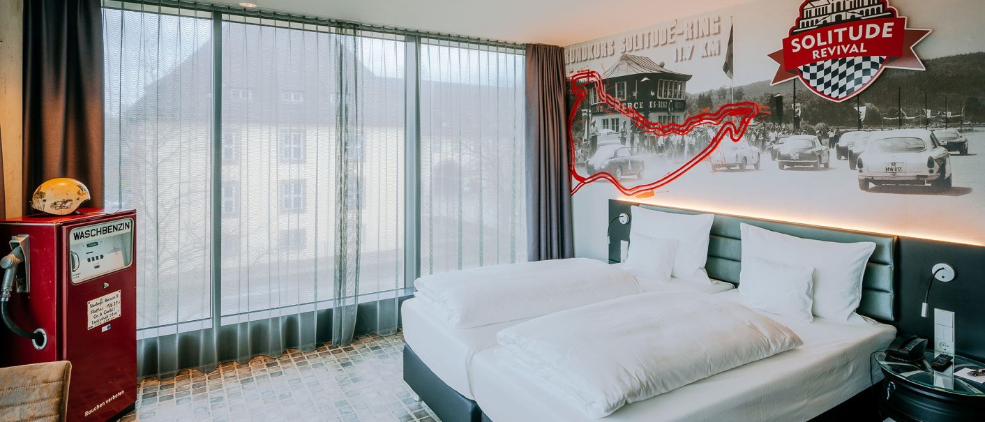 Hotelzimmer im V8 HOTEL mit Rennstrecken-Dekor, Doppelbett, Retro-Tankstelle und großem Fenster. An der Wand historische Rennfotos und Solitude Revival Logo., © V8 HOTEL Hotelzimmer im V8 HOTEL mit Rennstrecken-Dekor, Doppelbett, Retro-Tankstelle und großem Fenster. An der Wand historische Rennfotos und Solitude Revival Logo., © V8 HOTEL