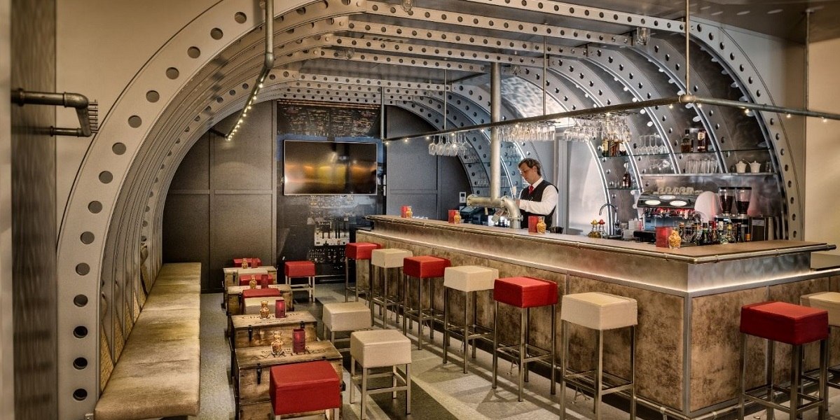 Moderne Bar mit industriellem Design, Metallbögen und Bartresen. Rote und weiße Hocker, gemütliche Sitzbereiche und ein Barkeeper hinter der Theke., © Essential by Dorint