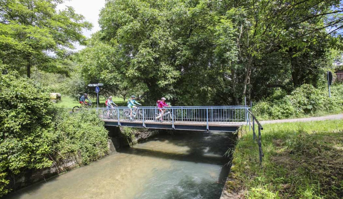 Radfahrer überqueren eine kleine Brücke über einen Fluss, umgeben von grünen Bäumen und Büschen., © Schwäbische Alb Tourismusverband e.V. Radfahrer überqueren eine kleine Brücke über einen Fluss, umgeben von grünen Bäumen und Büschen., © Schwäbische Alb Tourismusverband e.V.