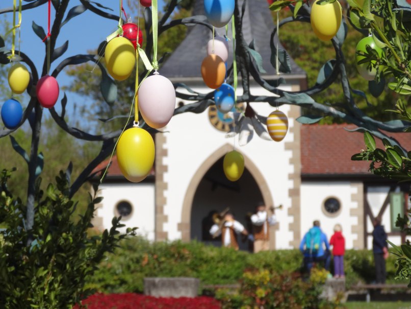 Bunte Ostereier h&auml;ngen an einem Baum, im Hintergrund ein Geb&auml;ude mit Menschen. Fr&uuml;hlingshafte Szene mit festlicher Dekoration., &copy; Erlebnispark Tripsdrill