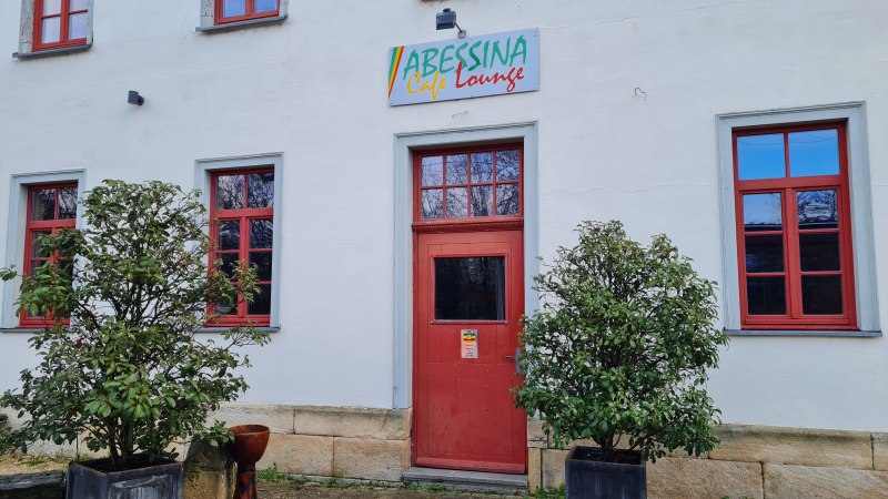 Der Eingangsbereich der Abessina Caf&eacute; Lounge mit roter T&uuml;r und Fenstern, flankiert von zwei Pflanzen in K&uuml;beln., &copy; Abessina