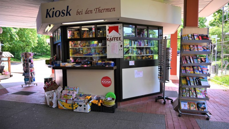 Ein Kiosk mit der Aufschrift 'Kiosk bei den Thermen', umgeben von Zeitschriftenst&auml;ndern und Verkaufsartikeln. Im Hintergrund sind B&auml;ume sichtbar.