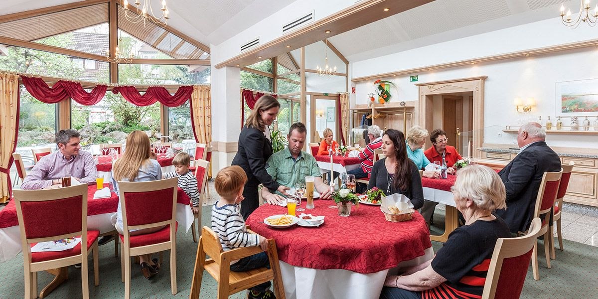 Ein Restaurant mit großen Fenstern, roter Tischdeko und mehreren Gästen, die an Tischen sitzen. Eine Kellnerin serviert Essen., © Hotel Restaurant Lamm