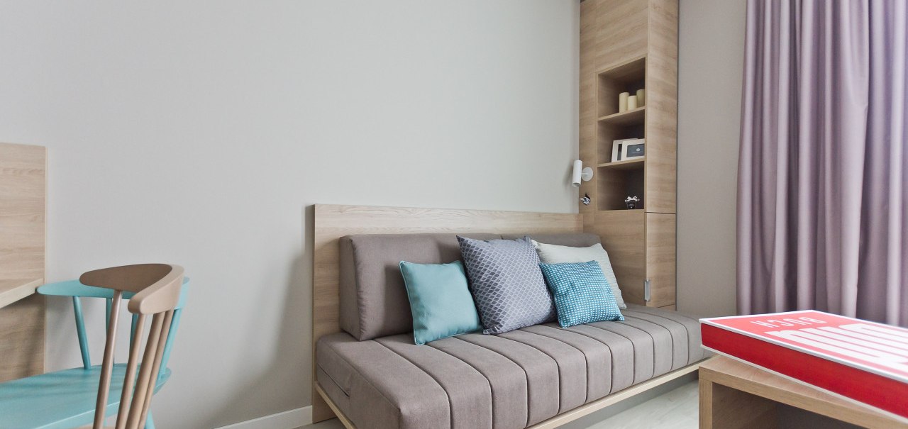 Modernes Apartment mit grauem Sofa, bunten Kissen, Holzmöbeln und lila Vorhängen. Ein Tisch mit rotem Buch ist im Vordergrund sichtbar., © Arthotel Ana / elaya