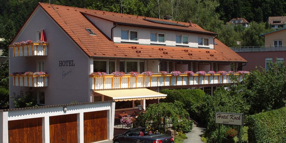 Hotel Koch mit roten Dachziegeln, blumengeschmückten Balkonen und einer Markise. Umgeben von grüner Vegetation und Garagen im Vordergrund., © Hotel Koch