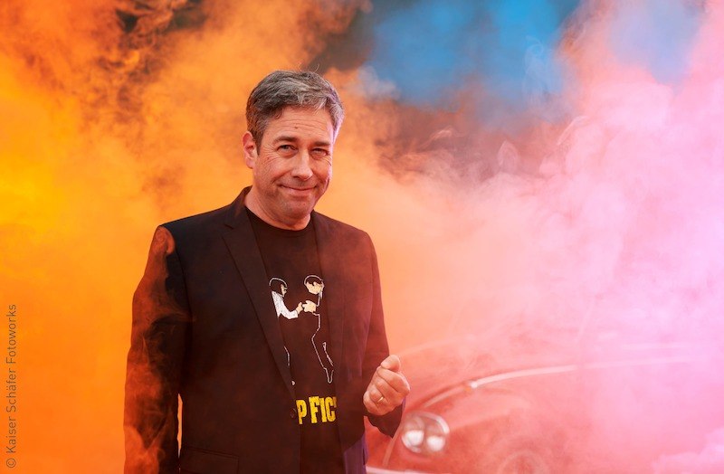 Ein Mann in einem schwarzen Anzug mit T-Shirt steht vor buntem Rauch in Orange und Blau. Im Hintergrund ist ein Auto zu erkennen., © Stadt Kornwestheim Kulturmanagement