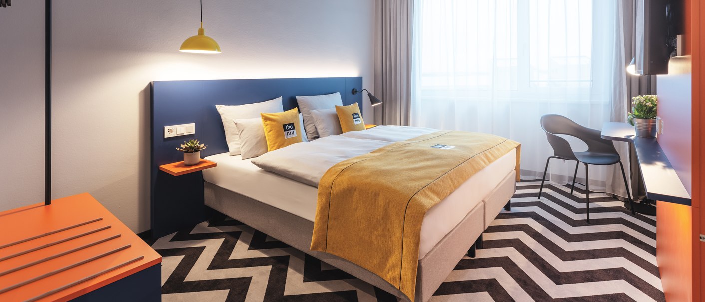 Modernes Hotelzimmer mit Doppelbett, gelber Bettdecke, blauem Kopfteil, Zickzack-Teppich und Schreibtisch mit Stuhl., © the niu Form - Novum Hospitality