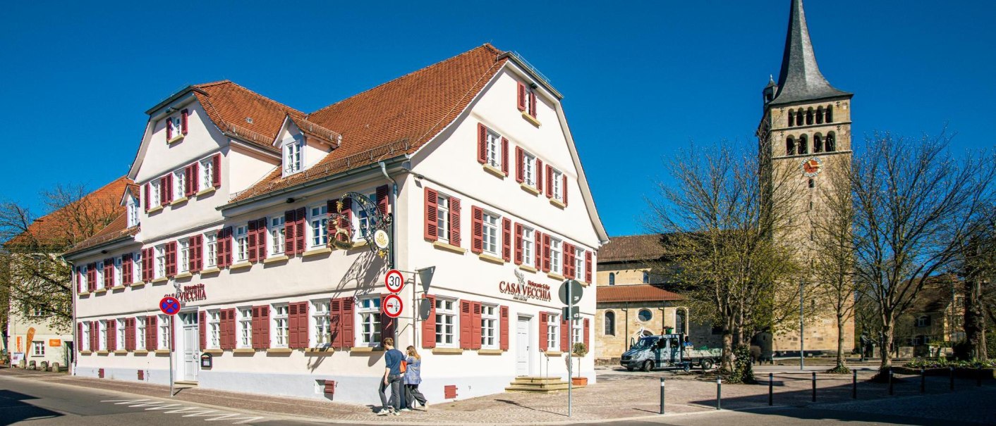 Ein historisches Gebäude mit roten Fensterläden in Sindelfingen, daneben ein Kirchturm. Menschen gehen auf dem Gehweg, blauer Himmel im Hintergrund., © Stuttgart-Marketing GmbH, Sarah Schmid Ein historisches Gebäude mit roten Fensterläden in Sindelfingen, daneben ein Kirchturm. Menschen gehen auf dem Gehweg, blauer Himmel im Hintergrund., © Stuttgart-Marketing GmbH, Sarah Schmid