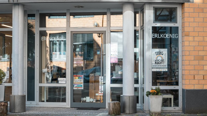 Eingang eines Gebäudes mit Glastüren, auf denen 'ERLKOENIG' steht. Poster und Pflanzen sind sichtbar. Moderne Architektur mit Backsteinfassade., © SMG, Sarah Schmid