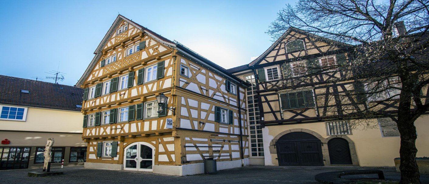 Fachwerkhäuser in Schorndorf, darunter das Stadtmuseum, bei sonnigem Wetter. Ein Baum und eine Skulptur sind ebenfalls zu sehen., © SMG, Sarah Schmid
